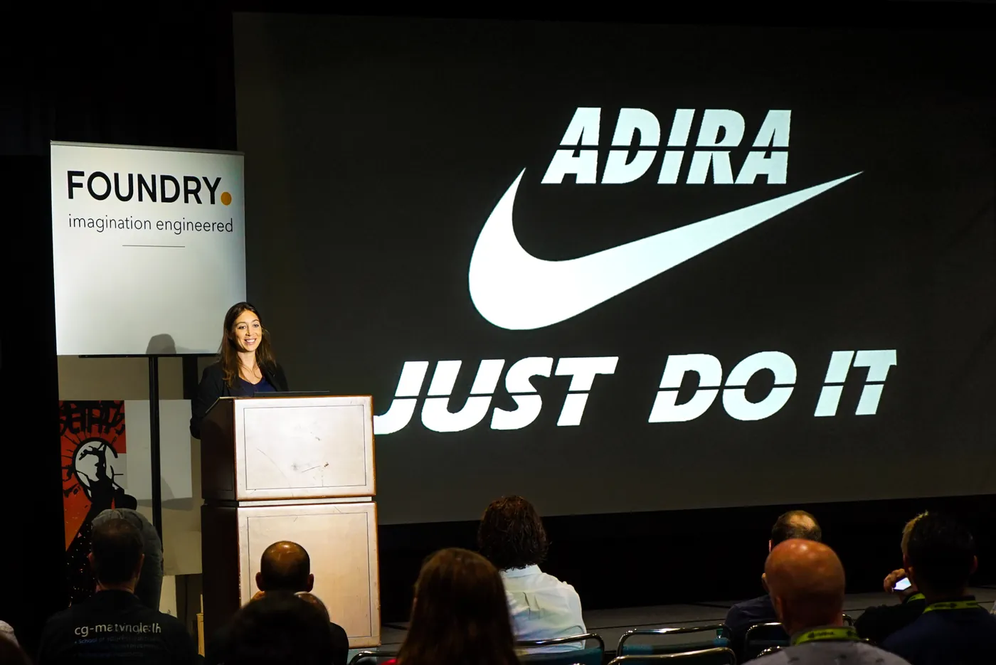 An Adira slide reading Just Do It, projected above the podium while an audience watches