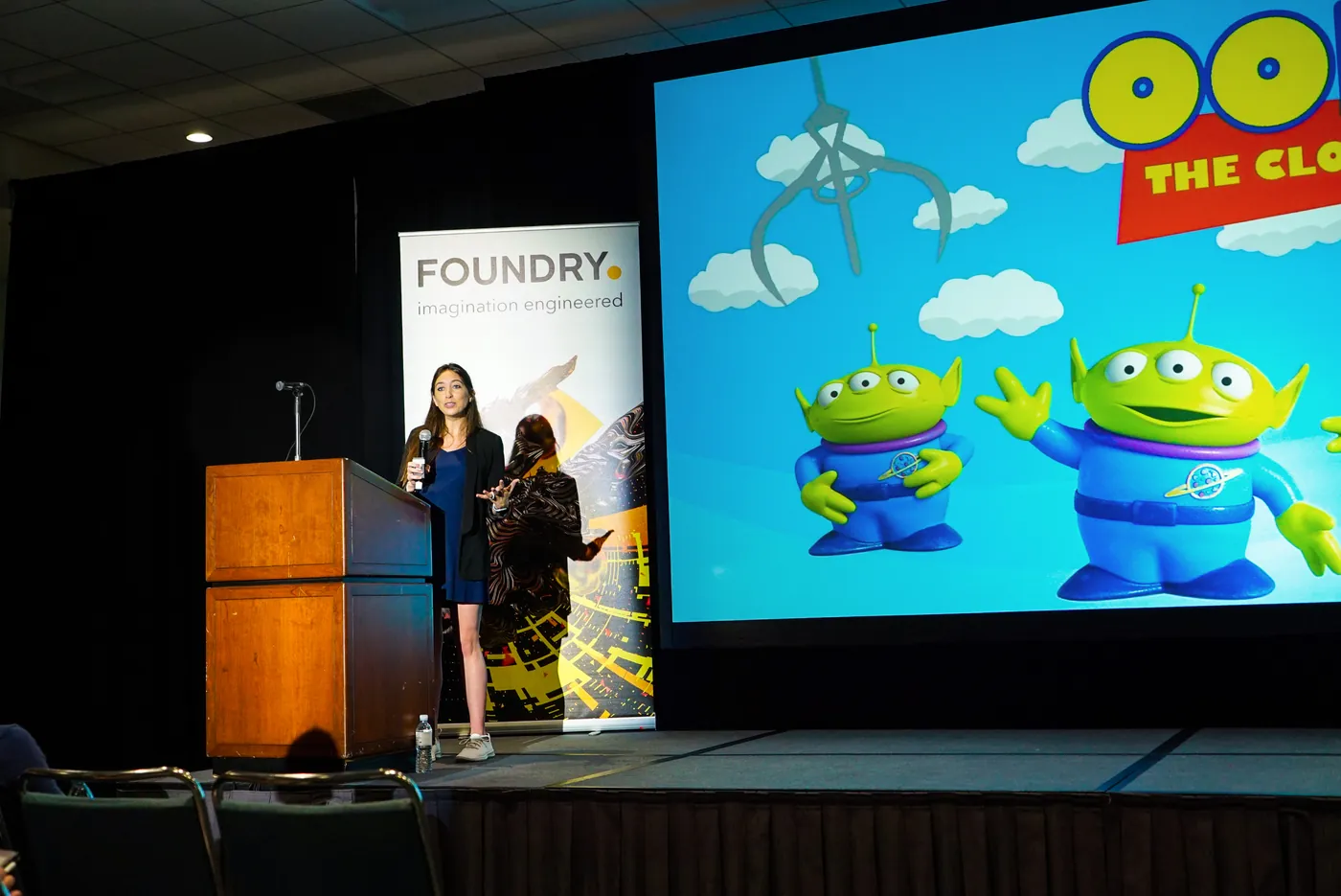 Two presenters on stage at the Foundry session in front of a large screen showing the Toy Story aliens